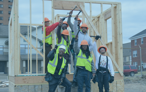 Former OIC affiliate students standing in front of the frame of a house. They're wearing orange hard hats, yellow vests, and holding their hammers up in the air.